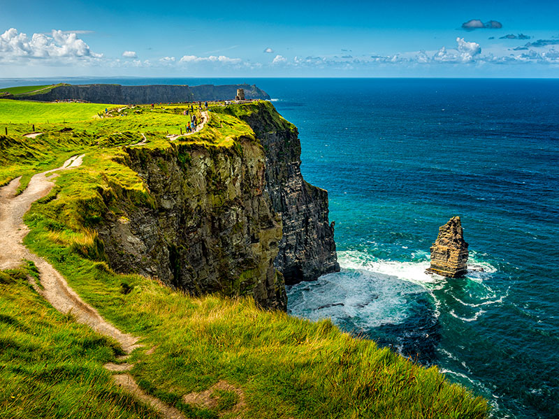 Lush green grasses top the jagged Cliffs of Moher on the west coast of Ireland near the village of Liscannor in County Clare, Republic of Ireland. Hikers make their way down the trail that hugs the cliff edge. The Atlantic Ocean stretches to the horizon. ©istockphoto by no_limit_pictures