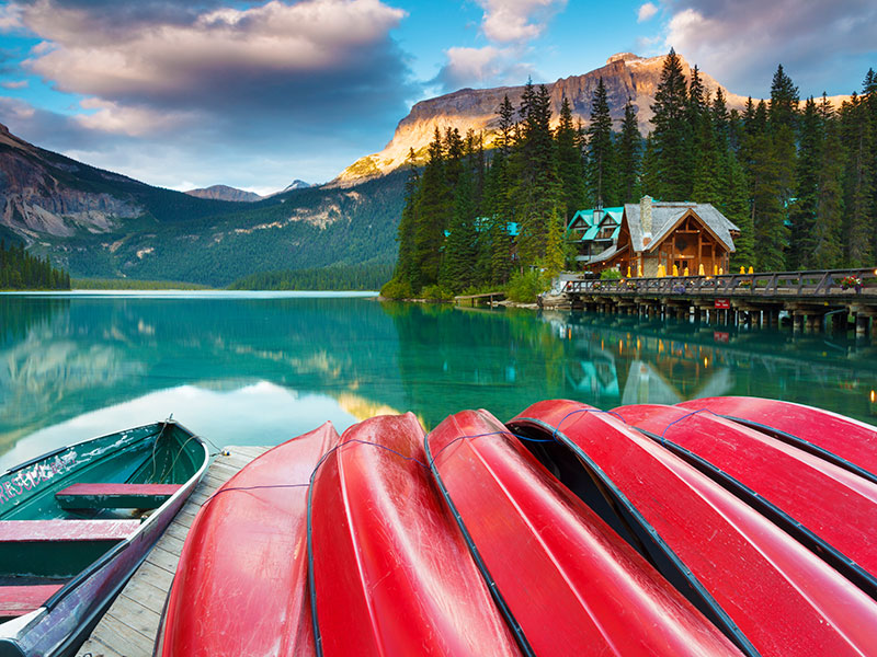 Emerald Lake at Yoho National Park, British Columbia, Canada. A line of red canoes is in the foreground, on a dock on the lake. In the background are the Rocky Mountains. To the left is a pier, a large wood timber building, and a grove of pine trees. ©istockphoto by GlowingEarth