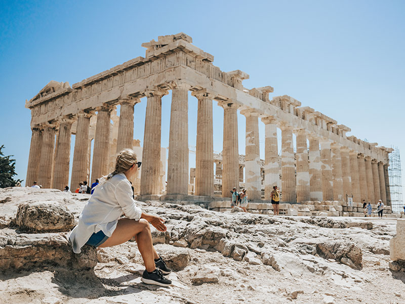 A woman, sitting in a rock in the foreground, looks a the ruins of the Parthenon, set against a bright, clear, blue sky. ©istockphoto by SimonSkafar
